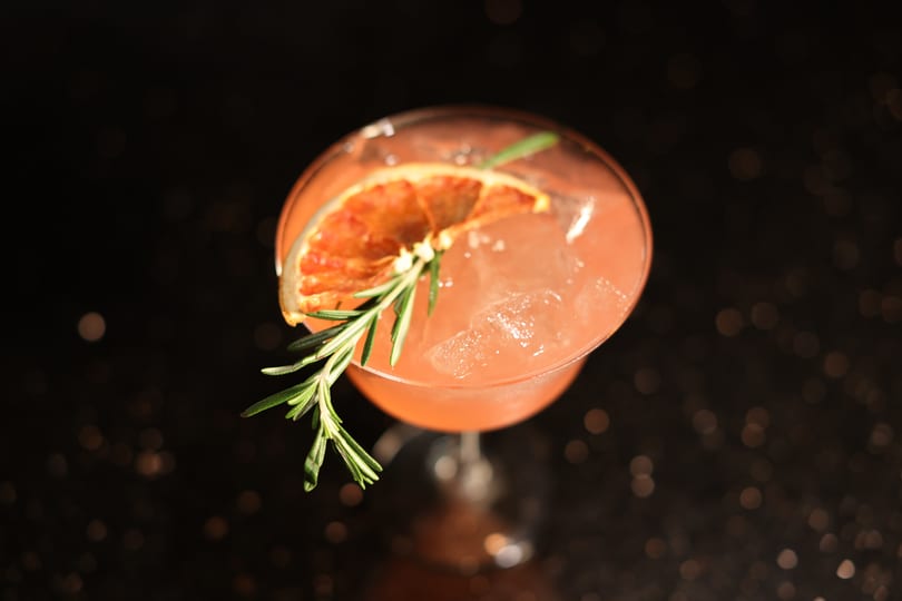 Pink cocktail with ice, garnished with a dried orange slice and a sprig of rosemary.
