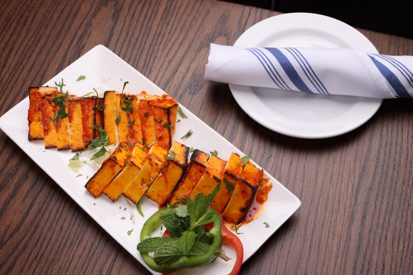 Grilled paneer tikka slices with red sauce and cilantro on a white platter, with a rolled napkin.