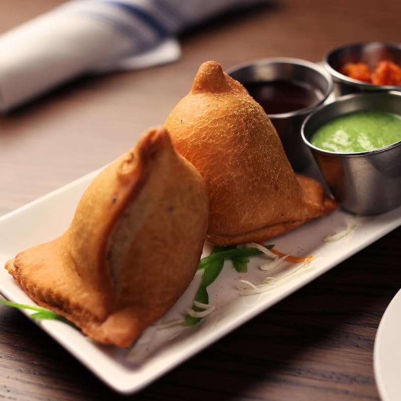 Two golden-brown samosas on a white plate with green and brown dipping sauces in metal cups.